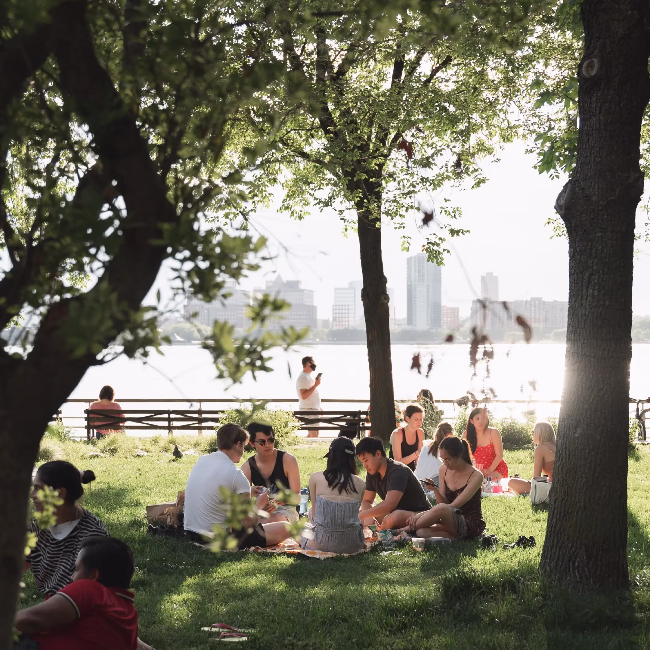 A group of college/university age students sitting on a blanket in the grass beside an urban river with a city skyline in the background.