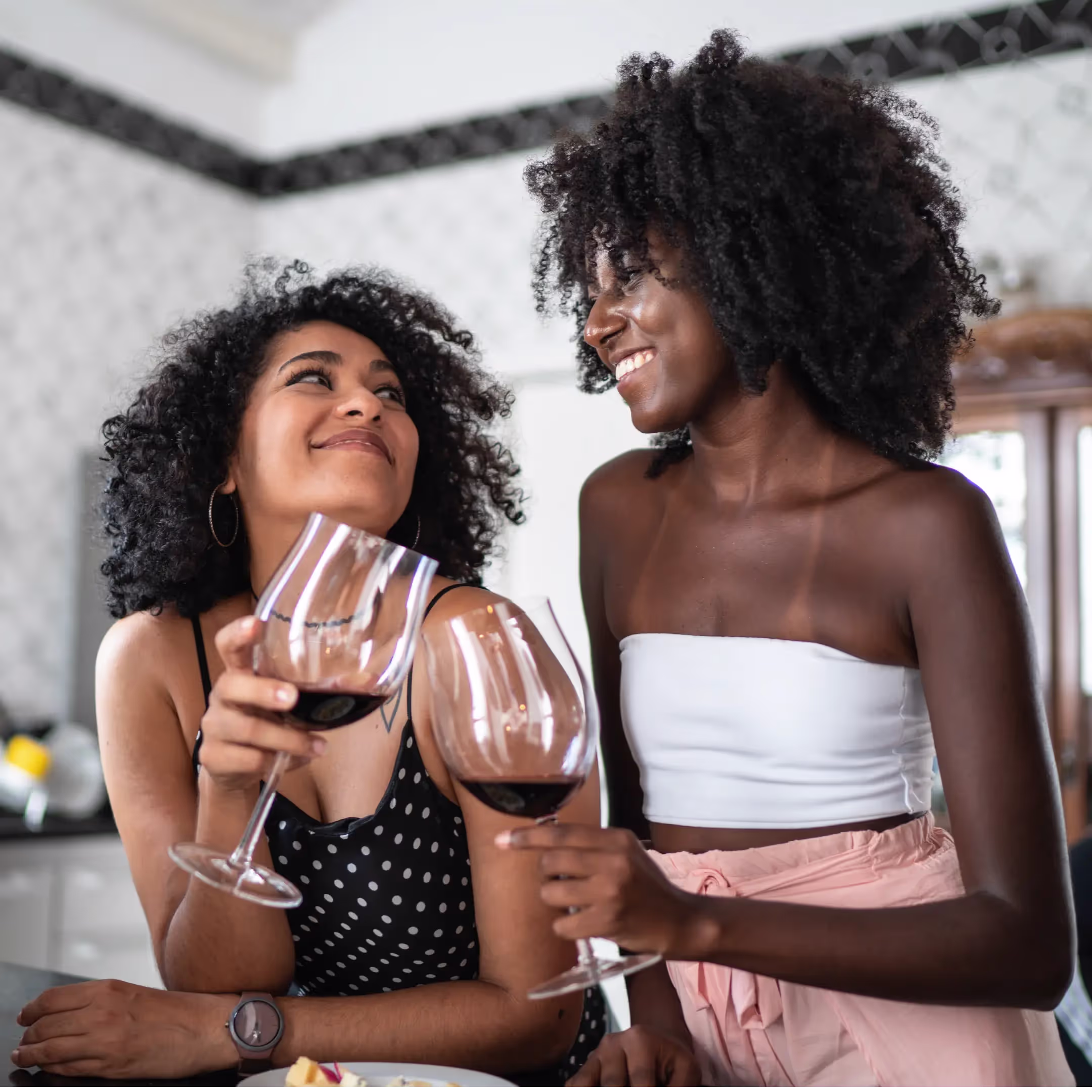 two black women standing in a kitchen, looking at each other smiling and clinking wine glasses together